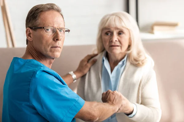 Selective focus of doctor in glasses holding hands with sick senior woman — Stock Photo