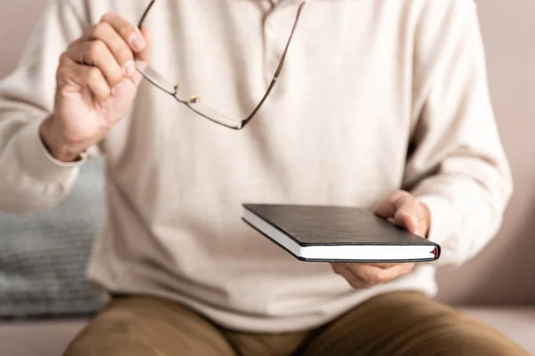 Vista recortada del anciano enfermo con dislexia sosteniendo gafas y cuaderno — Stock Photo