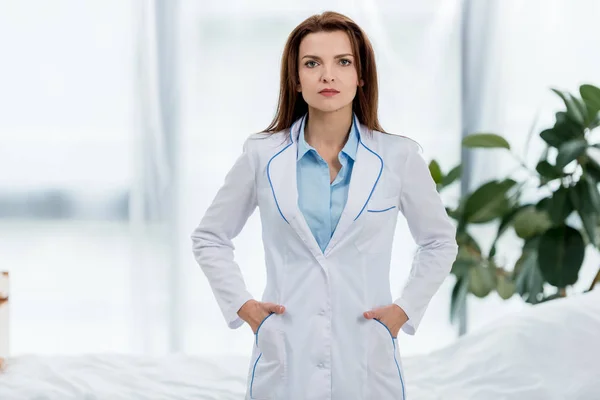 Attraktive Ärztin im weißen Mantel mit Händen in den Taschen, die im Krankenhaus in die Kamera schauen — Stockfoto