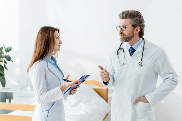 Médico de bata blanca mostrando el pulgar hacia arriba y hablando con su colega en el hospital - foto de stock