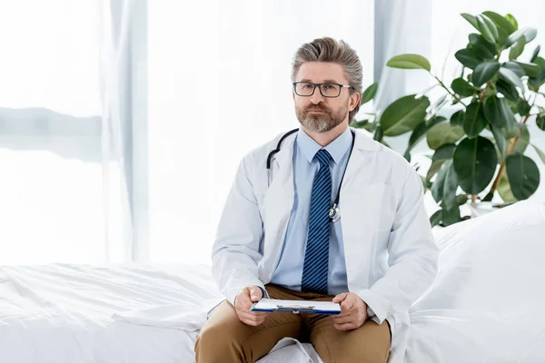 Handsome doctor in white coat sitting on bed and holding clipboard in hospital — Stock Photo