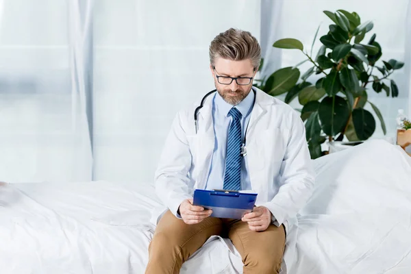 Schöner Arzt in weißem Mantel sitzt auf dem Bett und schaut im Krankenhaus auf Klemmbrett — Stockfoto