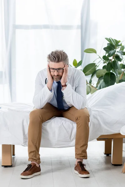 Sad doctor in white coat sitting on bed and looking down in hospital — Stock Photo