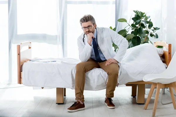 Nachdenklicher Arzt in weißem Mantel sitzt auf Bett und schaut im Krankenhaus weg — Stockfoto