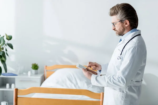 Vista laterale del bel medico in camice bianco con tablet digitale in ospedale — Foto stock