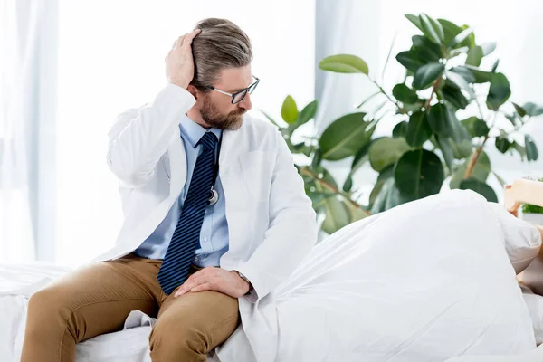 Schöner Arzt in weißem Mantel sitzt auf dem Bett und schaut im Krankenhaus weg — Stockfoto