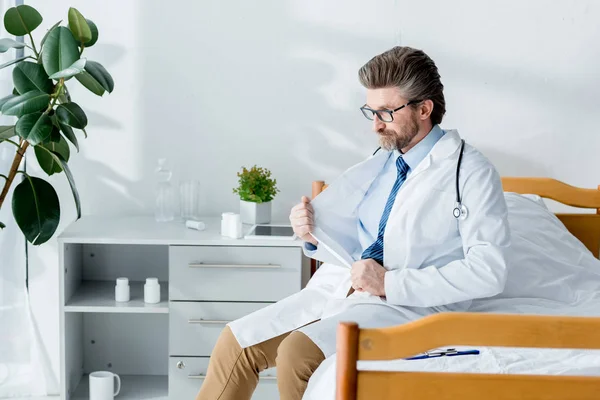 Bello medico in camice bianco seduto sul letto e guardando lontano in ospedale — Foto stock