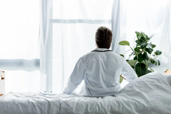 Rückenansicht eines Arztes im weißen Mantel, der im Krankenhaus auf dem Bett sitzt — Stockfoto