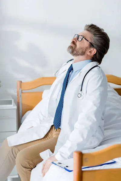 Handsome doctor in white coat sitting on bed and looking up in hospital — Stock Photo