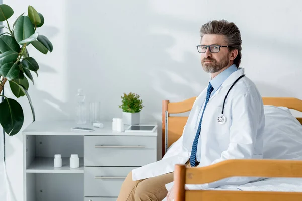 Handsome doctor in white coat sitting on bed and looking at camera in hospital — Stock Photo