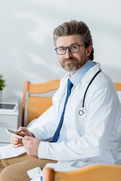 Beau médecin en manteau blanc assis sur le lit et tenant smartphone à l'hôpital — Photo de stock