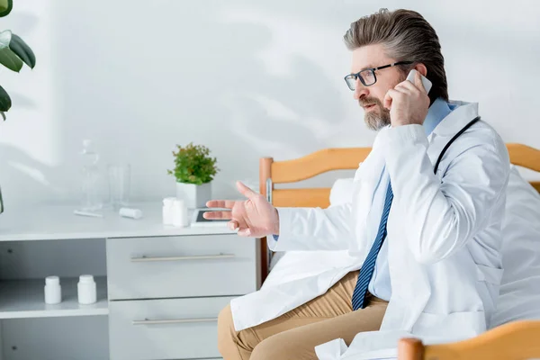 Schöner Arzt in weißem Mantel, der auf dem Smartphone spricht und im Krankenhaus auf dem Bett sitzt — Stockfoto