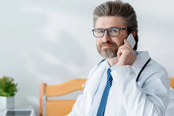 Guapo doctor en bata blanca hablando en teléfono inteligente en el hospital - foto de stock