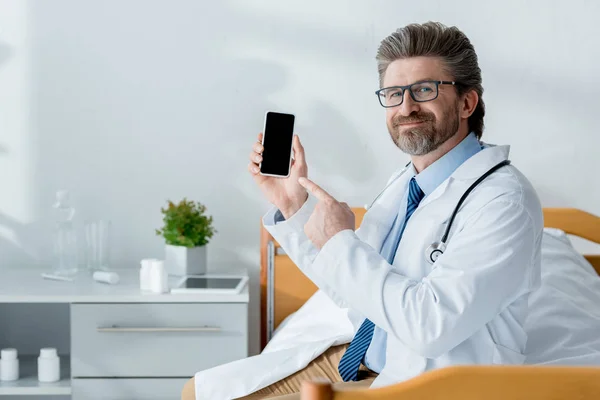 Médecin souriant en manteau blanc assis sur le lit et pointant du doigt le smartphone à l'hôpital — Photo de stock