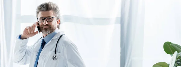 Panoramic shot of doctor in white coat talking on smartphone in hospital — Stock Photo