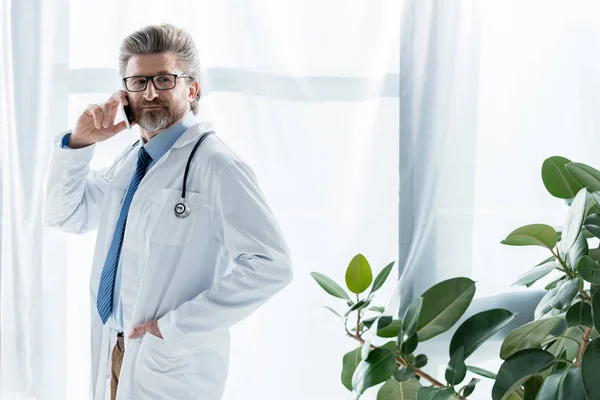 Handsome doctor in white coat with hand in pocket talking on smartphone in hospital — Stock Photo