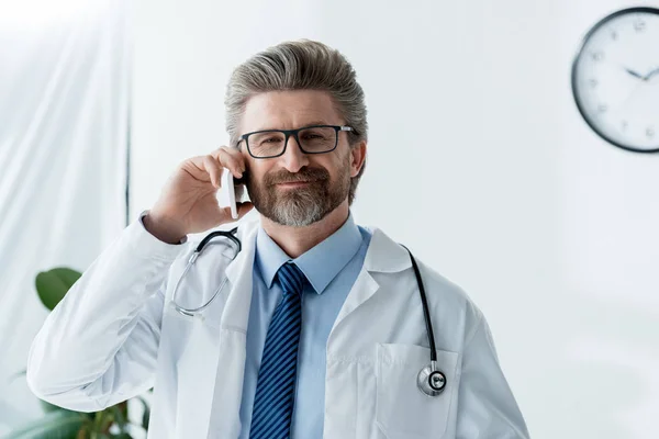 Улыбающийся доктор в белом халате разговаривает на смартфоне в больнице — стоковое фото