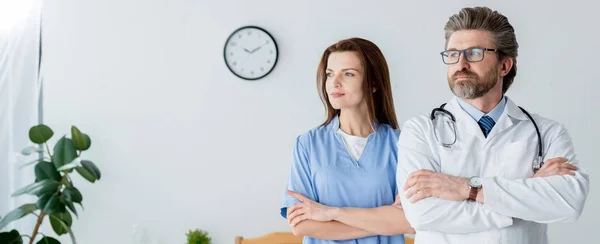 Панорамный снимок врача в белом халате и привлекательная медсестра со скрещенными руками, смотрящая в больницу — стоковое фото
