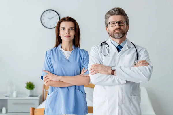 Doctor en bata blanca y atractiva enfermera con brazos cruzados mirando a la cámara en el hospital — Stock Photo