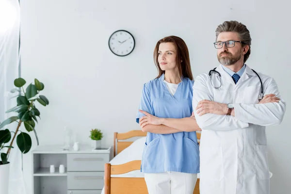 Лікар в білому пальто і приваблива медсестра з схрещеними руками дивиться далеко в лікарню — стокове фото