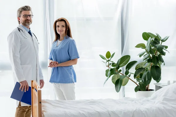 Médico sonriente en bata blanca y atractiva enfermera mirando a la cámara en el hospital - foto de stock