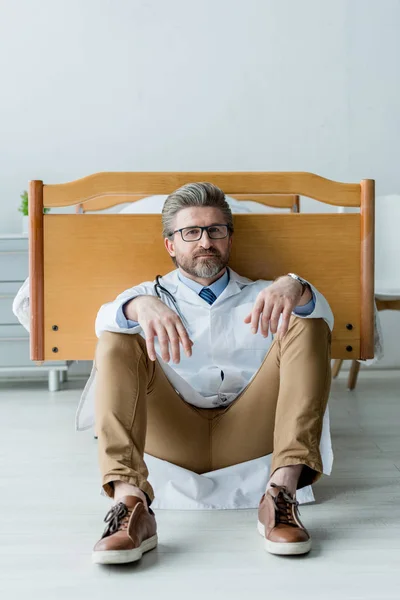Bello medico in camice bianco seduto sul pavimento e guardando la fotocamera in ospedale — Foto stock