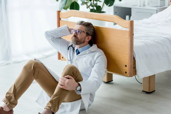Médico de bata blanca sentado en el suelo y tocando la cabeza en el hospital - foto de stock