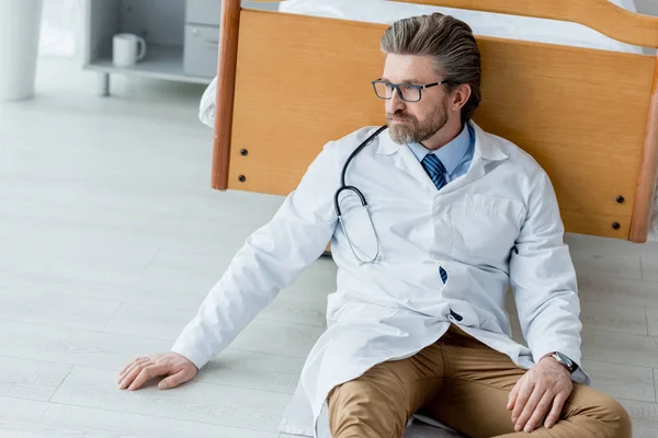 Schöner Arzt in weißem Mantel sitzt auf dem Boden und schaut im Krankenhaus weg — Stockfoto