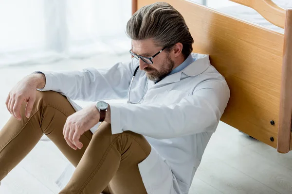 Handsome doctor in white coat sitting on floor and looking down in hospital — Stock Photo