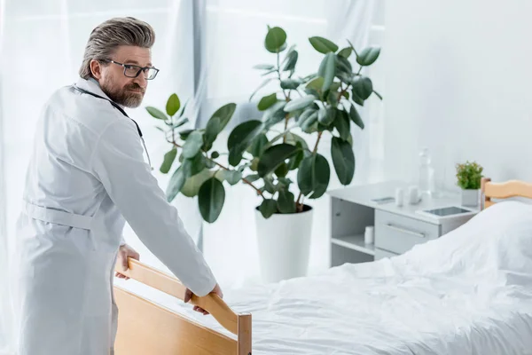 Schöner Arzt im weißen Mantel, der im Krankenhaus das Bett berührt und in die Kamera schaut — Stockfoto