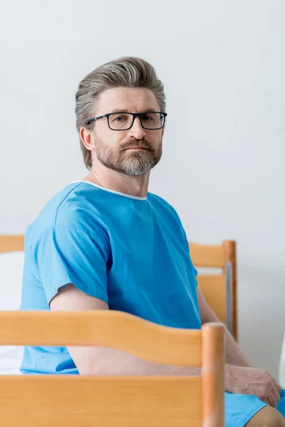 Patient im Arztkittel sitzt auf Bett und blickt im Krankenhaus in die Kamera — Stockfoto