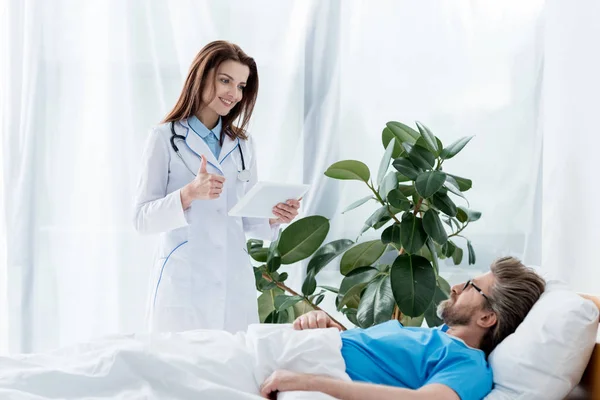 Medico sorridente in camice bianco che mostra come e tenendo in mano tablet digitale e paziente sdraiato a letto in ospedale — Foto stock