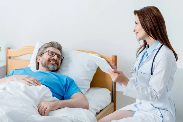 Médecin souriant en manteau blanc tenant comprimé numérique et regardant le patient au lit à l'hôpital — Photo de stock