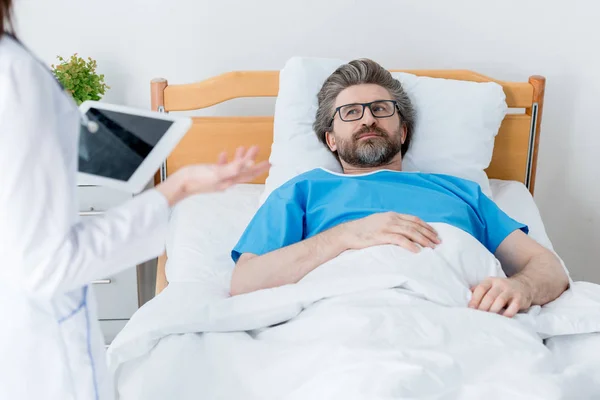 Vue recadrée du médecin tenant la tablette numérique et du patient couché au lit à l'hôpital — Photo de stock