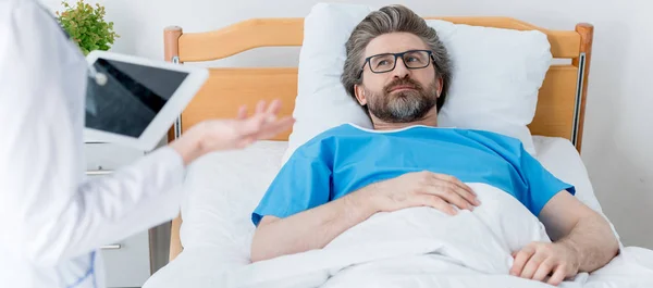 Inyección panorámica de médico sosteniendo tableta digital y paciente acostado en la cama en el hospital - foto de stock