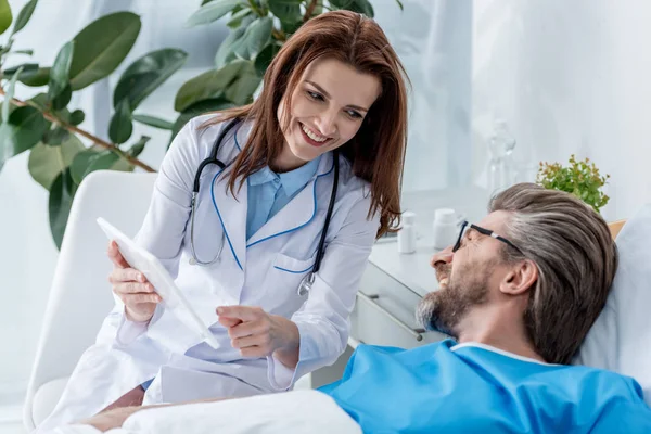 Médico sonriente de bata blanca apuntando con el dedo a la tableta digital y mirando al paciente - foto de stock