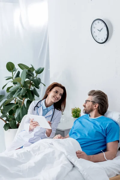 Улыбающийся доктор в белом халате, держащий цифровой планшет и смотрящий на пациента — стоковое фото