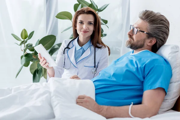 Médico sonriente de capa blanca sosteniendo tableta digital y paciente acostado en la cama en el hospital - foto de stock