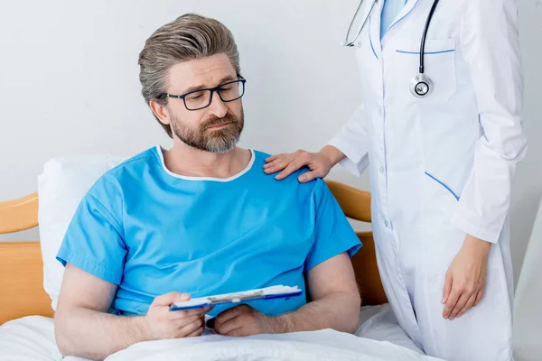 Cropped view of doctor in white coat touching shoulder of sad patient with clipboard — Stock Photo