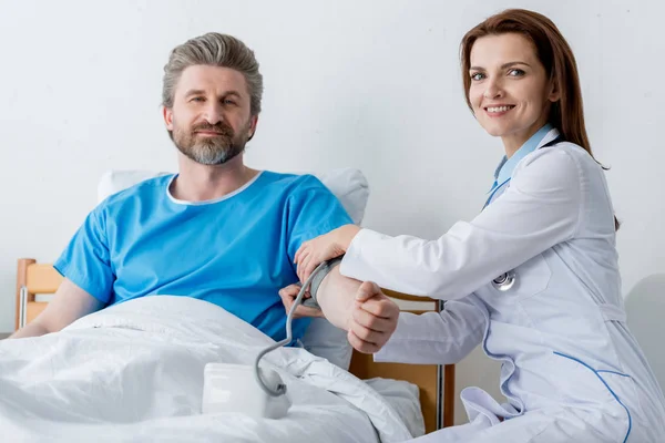 Médico sonriente de bata blanca midiendo la presión arterial del paciente en el hospital - foto de stock