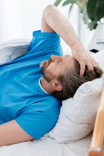 Paciente em vestido médico deitado e tocando a cabeça no hospital — Fotografia de Stock