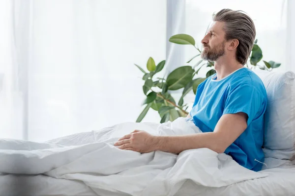 Side view of patient in medical gown sitting on bed in hospital — Stock Photo
