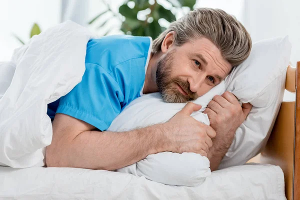 Красивый пациент в медицинском халате лежит в постели и смотрит в сторону — стоковое фото