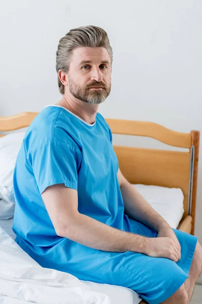 Paciente en bata médica sentado en la cama y mirando a la cámara en el hospital - foto de stock