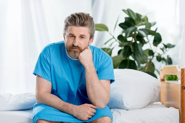 Pensive patient in medical gown sitting on bed in hospital — Stock Photo