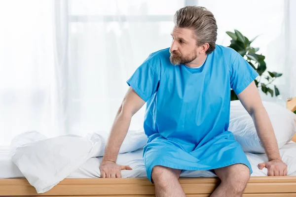 Patient en robe médicale assis sur le lit et regardant loin à l'hôpital — Photo de stock