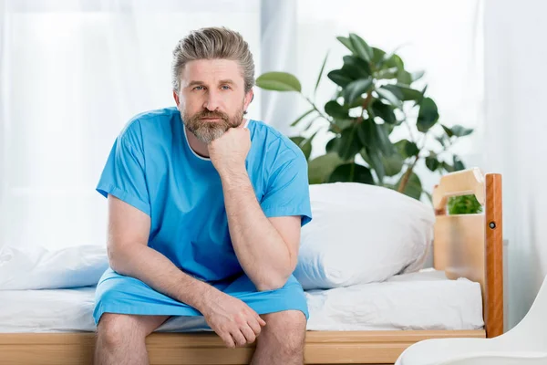 Nachdenklicher Patient im Arztkittel sitzt im Krankenhaus auf Bett — Stockfoto