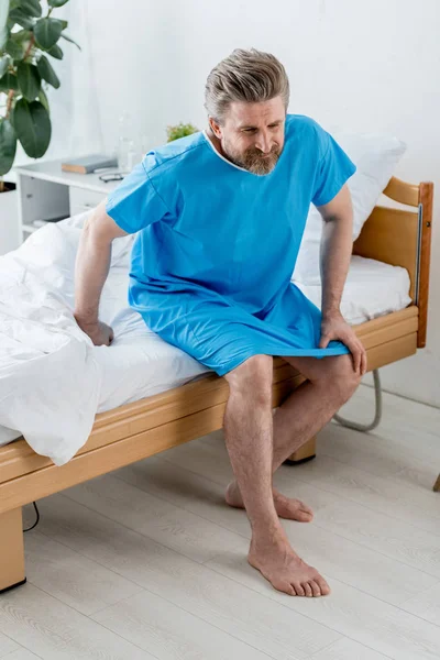 Vista de ángulo alto del paciente en bata médica sentado en la cama y sintiendo dolor de rodilla en el hospital - foto de stock