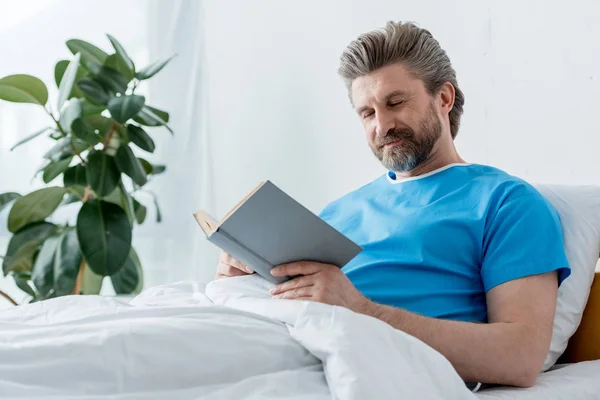 Guapo paciente en vestido médico libro de lectura en el hospital - foto de stock