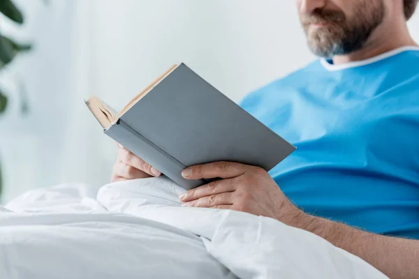 Обрезанный вид пациента в медицинском халате книги чтения в больнице — стоковое фото